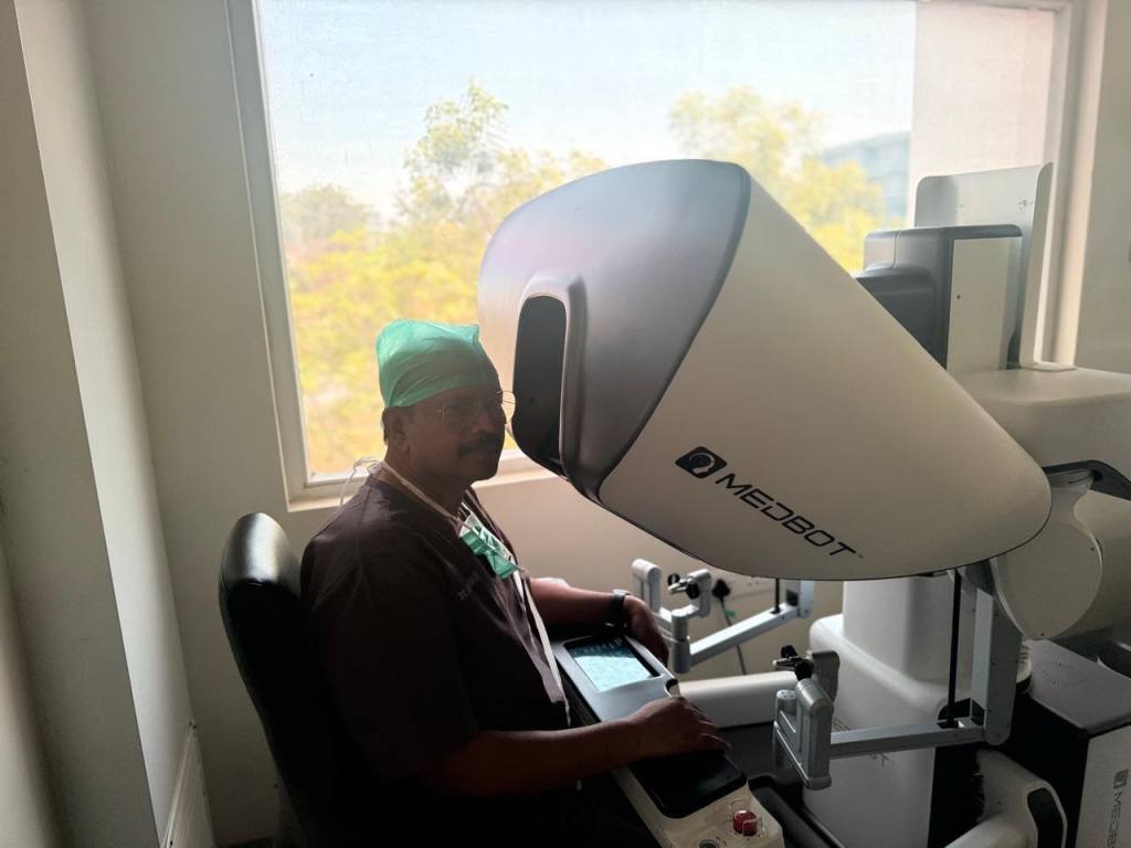 Surgeon wearing a green surgical cap and glasses sits at a Meubot robotic surgery console by a windowed room, operating the controls.